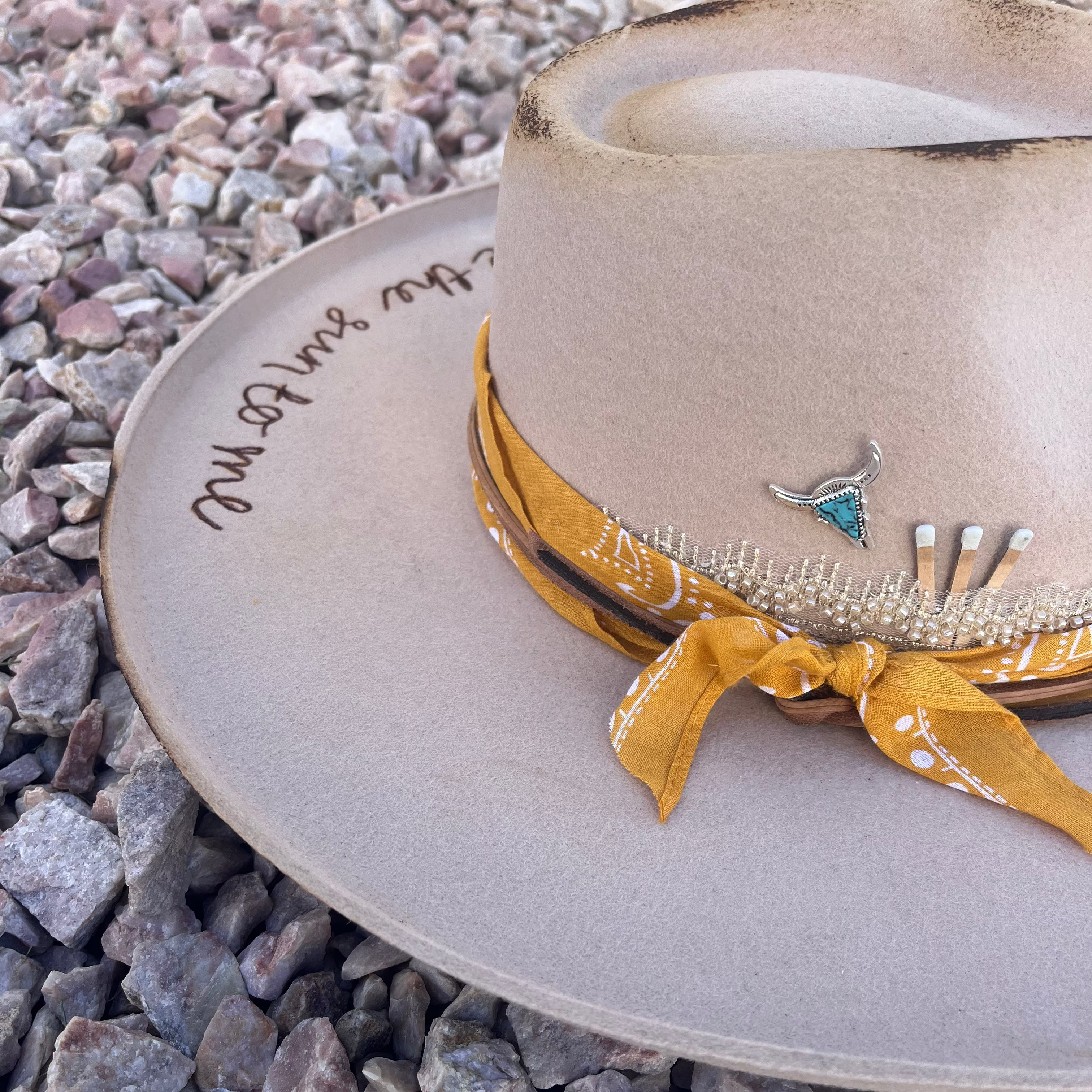A wide-brimmed cowboy hat in a light beige/tan color, styled outdoors on a bed of gray and white gravel. The hat features a yellow bandana-style ribbon with white paisley details, a small silver-toned longhorn pin with a turquoise accent, and black script lettering on the brim.