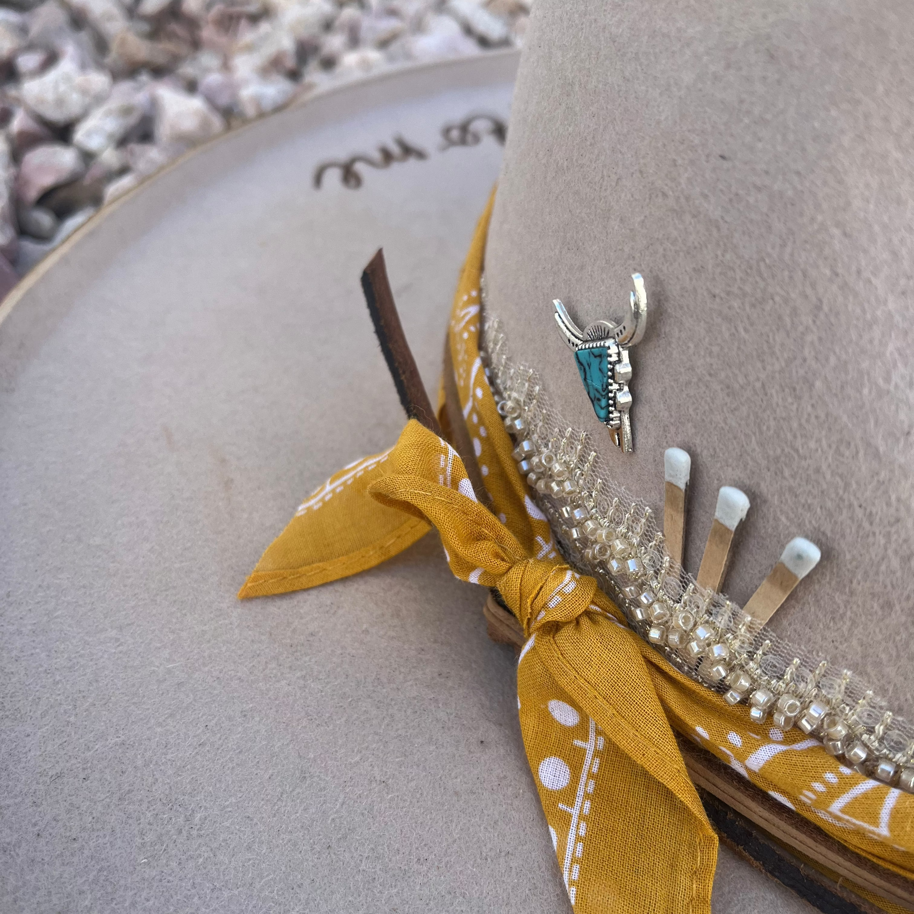 A close-up of a beige wide-brim hat with a mustard yellow bandana bow, rhinestone trim, and a silver cowboy badge, set against a background of small white shells.
